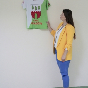 Pani Dyrektor Paulina Frontczak-Pawłowska trzyma powieszoną na ścianie koszulkę z logiem Gminy Grabów.