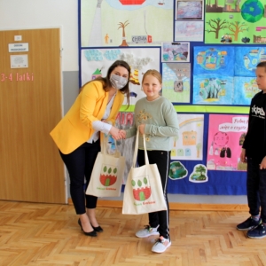 Pani Dyrektor Paulina Frontczak-Pawłowska uśmiech się do kamer razem z dziewczynką i trzymają torby z logiem Gminy Grabów, w tle kolorowe prace plastyczne.