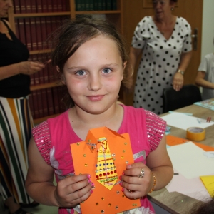 Uśmiechnięta dziewczynka pokazuje swoją pracę plastyczną w kształcie garnituru do kamery.