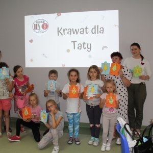 Grupowe zdjęcie dzieci które pokazują swoje prace na dzień ojca i Pani Dyrektor Paulina Frontczak-Pawłowska a nad nimi napis „Krawat dla Taty" i logo GCKB Grabów wyświetlane projektorem.
