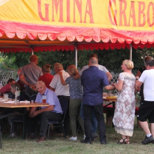 Stoły zapełnione gośćmi na Wiejskim Pikniku Rodzinnym w Byszewie, pod żółtym namiotem oznaczonym Gmina Grabów.