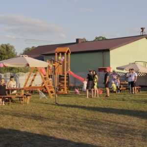 Grupka gości na Wiejskim Pikniku Rodzinnym w Byszewie., podczas jedzenia przy stolikach i na terenie placu zabaw.