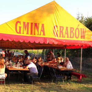 Stoły zapełnione gośćmi na Wiejskim Pikniku Rodzinnym w Byszewie, pod żółtym namiotem oznaczonym Gmina Grabów.
