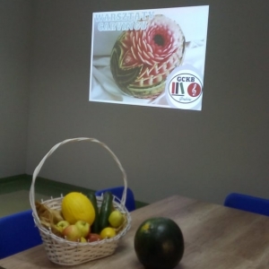 Stół z koszem z owocami i napis na ścianie „Warsztaty Carvingu" i logo GCKB Grabów wyświetlane projetktorem.