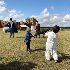 Dwóch uśmiechniętych chłopców w strojach średniowiecznych walczą ze sobą drewnianymi atrapami mieczy, a w tle widać kilku aktorów i ruiny zamku.