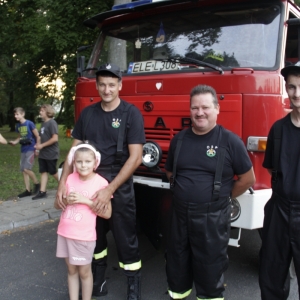 Trzech strażaków  z dziewczynką na tle wozu strażackiego.