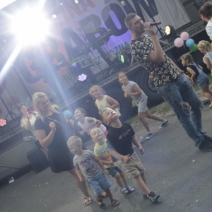 Grupka dzieci bawiąca się przed sceną wraz ze śpiewającym artystą wśród nich. 