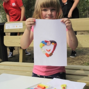 Uśmiechnięta dziewczynka pokazuje swój rysunek na kartce papieru z plasteliny przedstawiający uśmiechnięte serce.