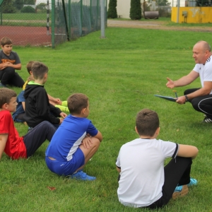 Dzieci siedzą na trawie i słuchają trenera.