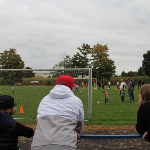 Dorośli i jeden chłopiec oglądają jak mały chłopiec kopie piłkę na boisku.
