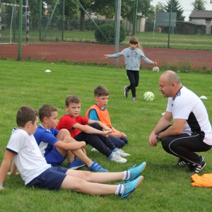 Dzieci siedzą na trawie i słuchają trenera.