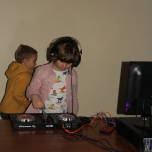 Jedno z dzieci stojące na stanowisku didżeja, ze słuchawkami na uszach ogląda sprzęt do odtwarzania muzyki.