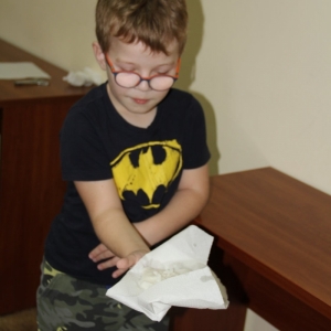 Zadowolony chłopiec w okularach i czarnej bluzce, trzyma na chusteczce swój wosk przelany przez klucz do wody. 