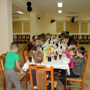 Długi stół zastawiony ciastkami, paluszkami, owocami, sokami i cukierkami, przy którym siedzą dzieci wraz z paniami opiekunkami.