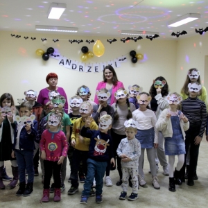 Zdjęcie grupowe dzieci w maskach w kształcie misia wraz z panią opiekunką i Panią Dyrektor Pauliną Frontczak-Pawłowską na tle napisu "Andrzejki".