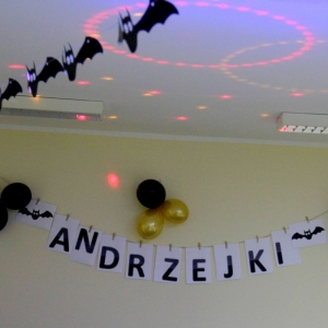 Dekoracja zawieszona na ścianie i pod sufitem, składająca się z napisu „Andrzejki", baloników czarnych i złotych oraz nietoperzy z papieru.