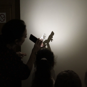 Grupka dzieci wraz z panią przyglądają się przy zgaszonym świetle i zapalonej latarce, jaki cień oddają na ścianie kształty powstałe z wosku.