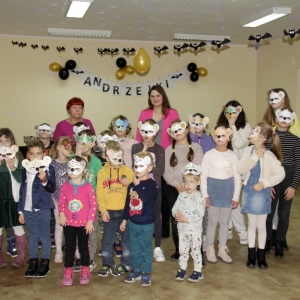 Zdjęcie grupowe dzieci w maskach w kształcie misia wraz z paniami opiekunkami na tle napisu "Andrzejki".