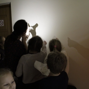 Grupka dzieci wraz z panią przyglądają się przy zgaszonym świetle i zapalonej latarce, jaki cień oddają na ścianie kształty powstałe z wosku po przelaniu go do wody.
