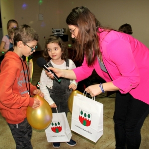 Pani Dyrektor Paulina Frontczak-Pawłowska wręcza chłopcu w okularach, torebkę z nagrodami, a chłopiec ten mówi do mikrofonu. Pozostała grupka dzieci czeka.