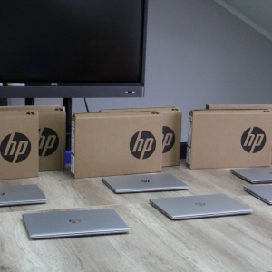 Sześć laptopów HP i opakowania do nich rozłożone na stole i monitor interaktywny postawiony za stołem.