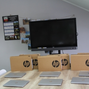 Sześć laptopów HP i opakowania do nich rozłożone na stole i monitor interaktywny postawiony za stołem.