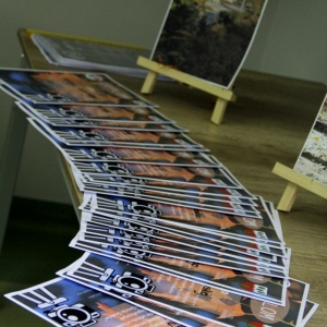 Komplet wypełnionych dyplomów zwycięzców konkursu rozłożone na stole wraz z wydrukowanymi zdjęciami leżącymi na podpórkach.  