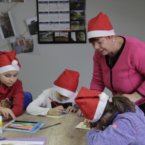 Pani prowadząca pomaga dzieciom  w czapkach Mikołaja robić kartki świąteczne.
