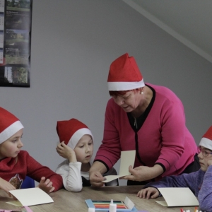 Pani prowadząca pomaga dzieciom  w czapkach Mikołaja robić kartki świąteczne.