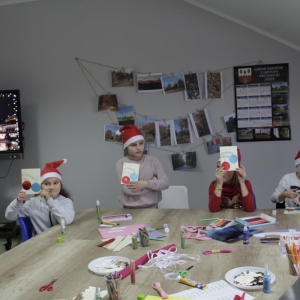 Dzieci z czapkami Mikołaja stoją przy stole z przyborami i pokazują zrobione przez siebie kartki świąteczne.