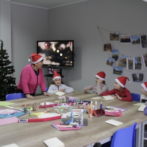Dzieci w czapkach Mikołaja dekoruję choinkę a w tle jest telewizor ze świąteczną składanką piosenek.