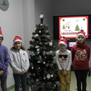 Dzieci w czapkach Mikołaja stoją przy udekorowanej choince a w tle jest telewizor ze świąteczną składanką piosenek.
