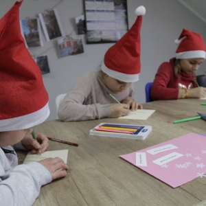 Dzieci z czapkami Mikołaja siedzą przy stole i robią kartki świąteczne.