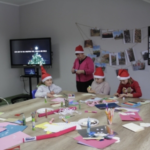 Dzieci z czapkami Mikołaja siedzą przy stole i robią kartki świąteczne a w tle jest telewizor ze świąteczną składanką piosenek.