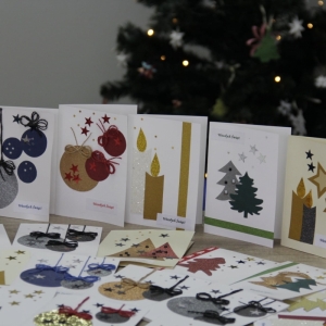 Własnoręcznie zrobione kartki świąteczne ustawione na stole a za nimi ozdobiona choinka.