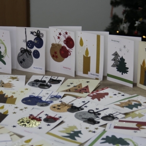 Własnoręcznie zrobione kartki świąteczne ustawione na stole a za nimi ozdobiona choinka.