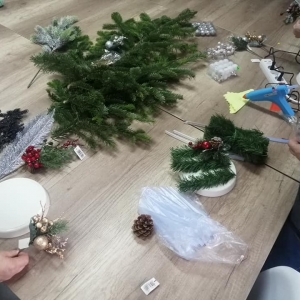 Uczestnicy stoją przy stole i używają przyrządów plastycznych, żeby zrobić stroiki świąteczne.