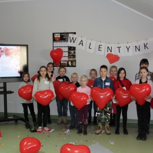 Dzieci stoją pod napisem „WALENTYNKI" i trzymają balony w kształcie serca.