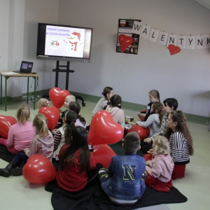 Dzieci siedzą z balonikami w kształcie serca i słuchają pani prowadzącej.