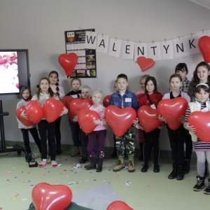 Dzieci stoją pod napisem „WALENTYNKI" i trzymają balony w kształcie serca.