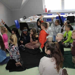 Dzieci siedzą i patrzą z zainteresowaniem, z czego niektóre z nich podnoszą rękę do góry, bo chcą coś powiedzieć.