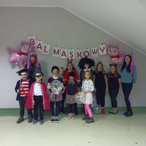 Różnie przebrane dzieci wraz z prowadzącymi stoją pod wywieszonym na ścianie napisem „BAL MASKOWY".