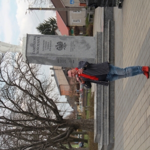 Pan prowadzący trzymający mikrofon na tle pomnika Tadeusza Kościuszki.