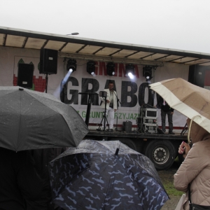 Pan prowadzący na scenie w Grabowie podczas pikniku Dzień Rodzinny i grupka słuchaczy z parasolami.