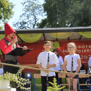 Dwójka dzieci na scenie trzymających instrumenty, a obok nich stoi przebrany za krasnala pan z mikrofonem.