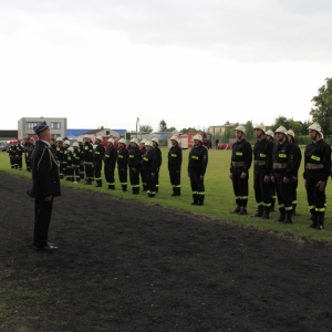 Strażacy w strojach służbowych stoją ustawieni w rzędzie na boisku, a przed nimi stoi strażak w stroju galowym.