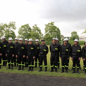 Grupa strażaków ubranych w strojach służbowych stoi w rzędzie na trawniku.