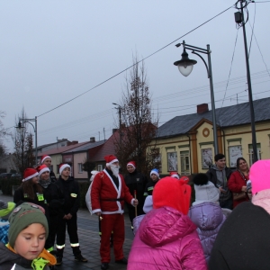 Mężczyzna przebrany za Świętego Mikołaja a za nim dzieci przebrane za strażaków.