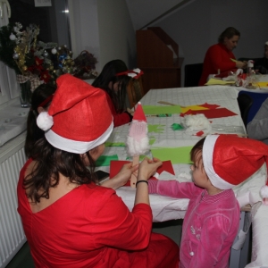 Dziewczynki w czapkach Mikołaja robią ozdoby świąteczne przy stołach.