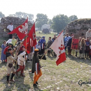 Grupa ludzi ucharakteryzowanych, część z nich trzyma flagi  i przedstawiają turniej rycerski na terenie z pozostałościami starego muru z kamieni i cegły. 